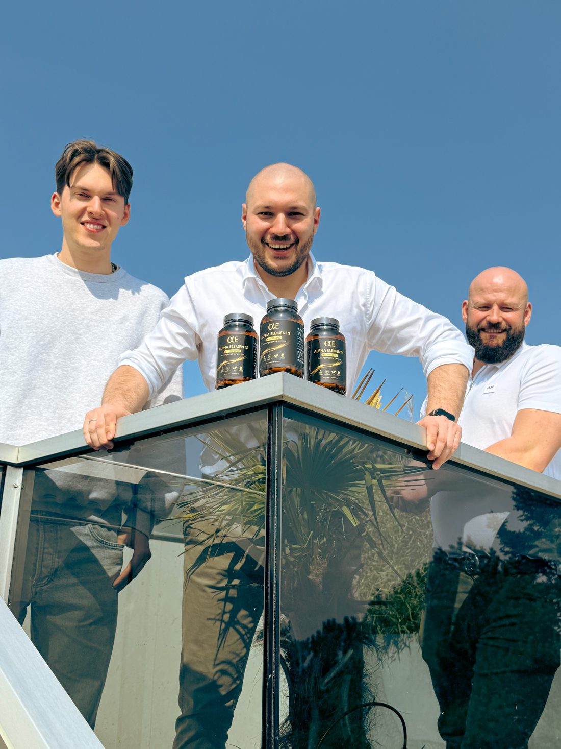 Gabriel, Darius und Thomas lehnen lachend an einem Glasgeländer, vor ihnen stehen drei Dosen ALL-IN-ONE
