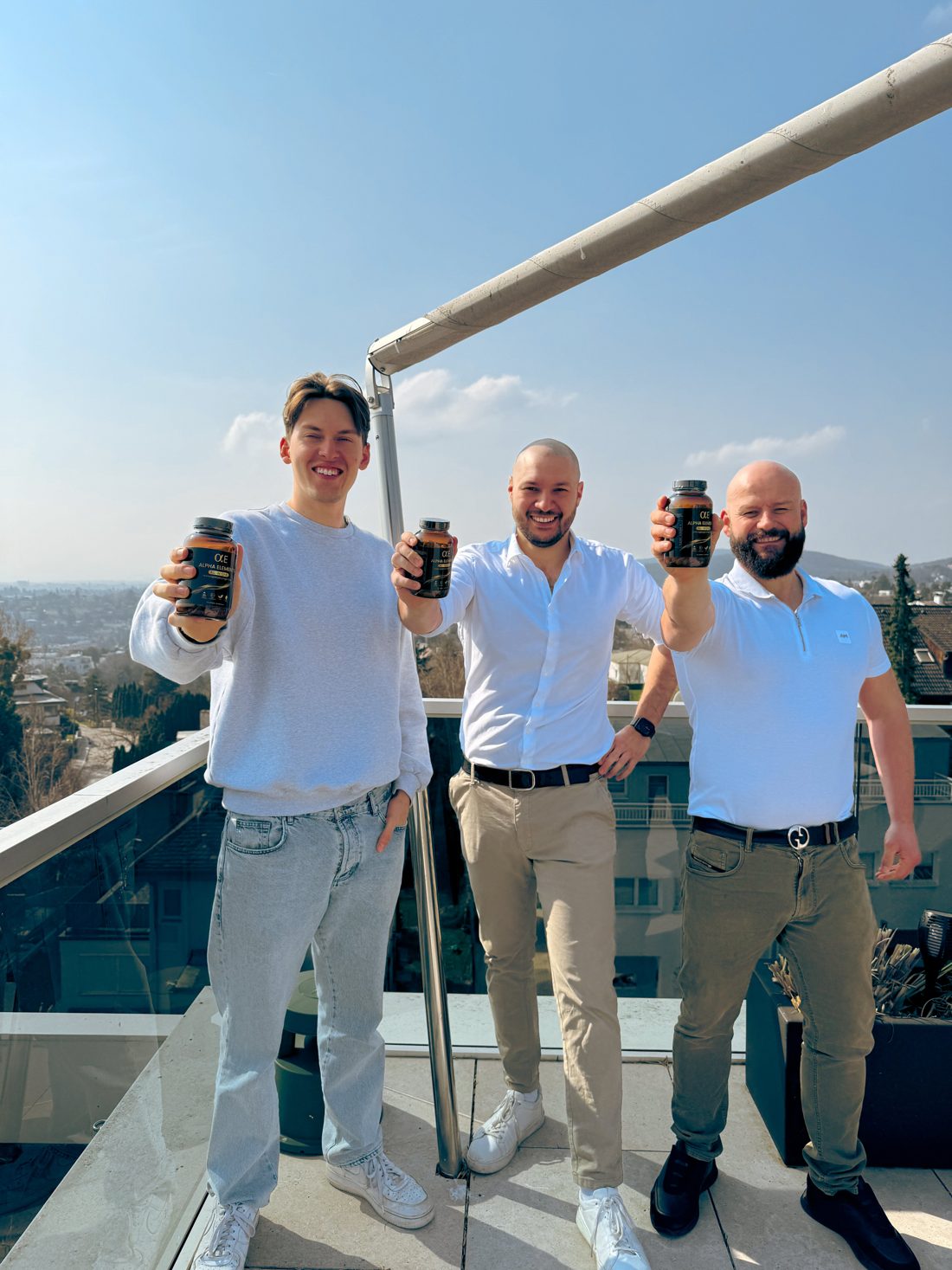 Die drei Gründer von Alpha Elements halten ALL-IN-ONE-Dosen in die Kamera, auf einer Terrasse mit Blick über Wien